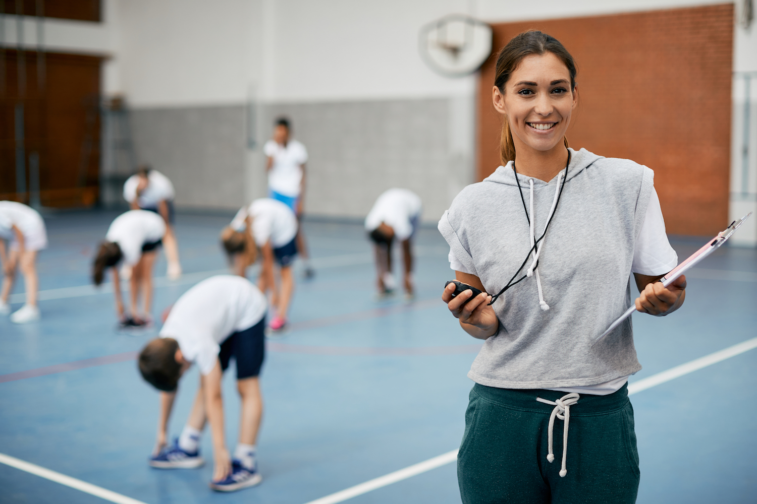 Young happy coach using stopwatch during PE class at school gym and looking at camera. Her students are exercising in the background.